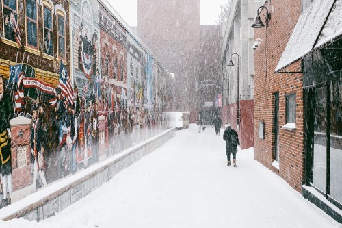 A snowy alleyway with a mural on one wall and a person walking through deep snow, surrounded by falling snowflakes.