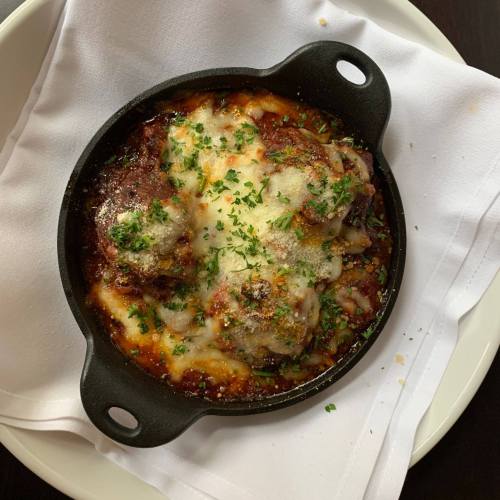A cast-iron dish of baked meatballs topped with melted cheese and herbs, served on a white napkin.
