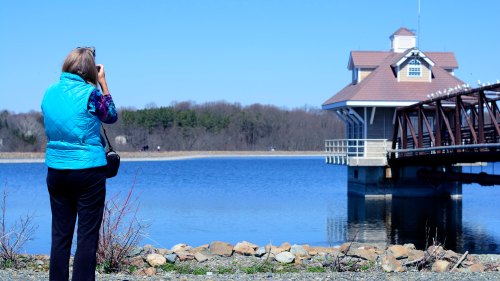 A person in a blue vest takes a photo of a lakeside building and bridge, surrounded by trees and water.
