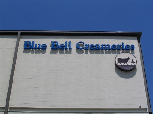 Sign for Blue Bell Creameries on a building, featuring a blue logo with a cow and a child. Clear blue sky above.