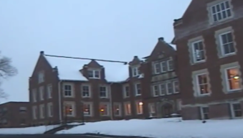 A historic brick building covered in snow, with large windows glowing warmly in the evening light.