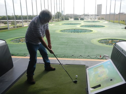 A person swings a golf club at a driving range with multiple target areas in the background.