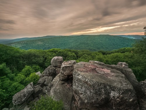 A rocky outcrop overlooks a lush green valley under a cloudy sky at sunset.
