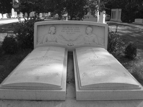 A black and white image of a double gravestone featuring two figures shaking hands, surrounded by greenery.