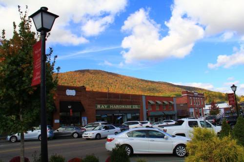 A small town street with shops, cars parked, and a backdrop of colorful autumn mountains under a blue sky.