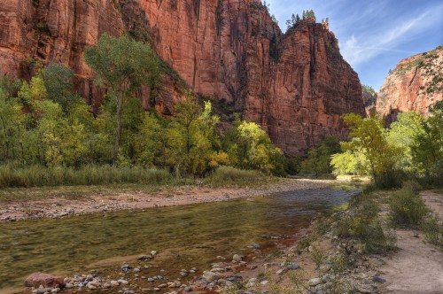 A serene river flows through a canyon with towering red cliffs and vibrant green trees under a blue sky.