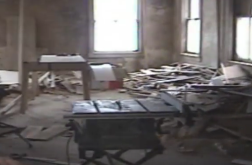 A cluttered, dusty room with a table saw and debris scattered on the floor, illuminated by natural light from windows.