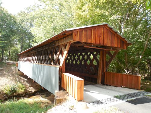 A wooden covered bridge with a triangular lattice design, surrounded by trees and a small creek.