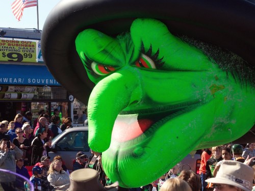 A large green witch balloon with a prominent nose and hat, looming over a crowd at a festive event.
