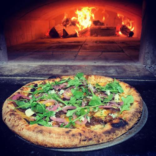 A freshly baked pizza topped with greens and meats, with a wood-fired oven glowing in the background.