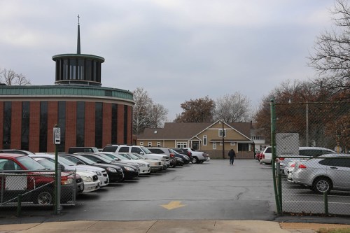 A parking lot with various cars, a church-like building, and a house in the background under a cloudy sky.