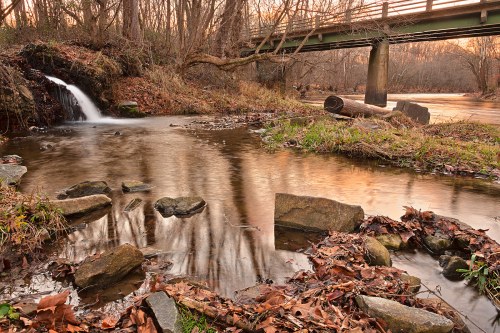 A serene landscape featuring a small waterfall, a stream, and a bridge surrounded by trees and fallen leaves.