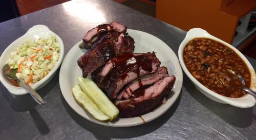 A plate of sliced barbecue meat with sauce, served with coleslaw, baked beans, and pickles on the side.