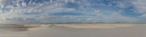 A panoramic view of a vast white sand desert under a blue sky with scattered clouds and distant mountains.