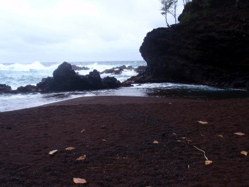 A rocky coastline with dark sand and waves crashing against the shore under a cloudy sky.