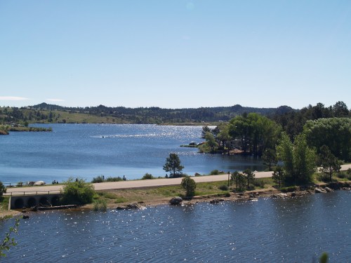 A serene lake surrounded by trees and hills, with sunlight reflecting on the water and a path along the shore.