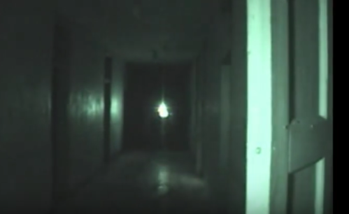 A dimly lit hallway with shadows, featuring a faint light source at the far end.