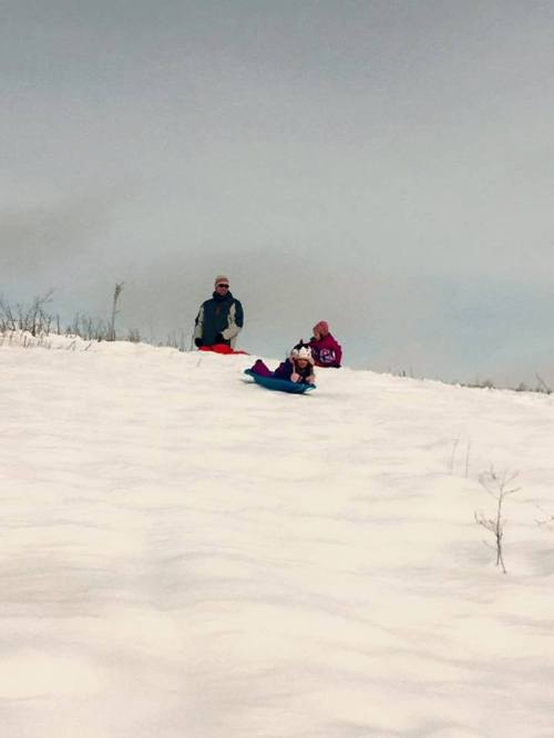 A snowy hill with two people, one sitting and one lying on a sled, enjoying a winter day.