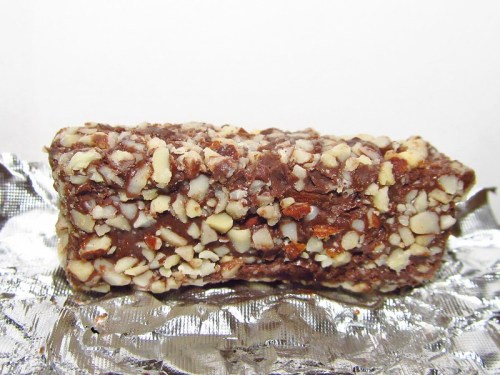 A chocolate-covered candy bar coated in chopped nuts, resting on a silver wrapper.