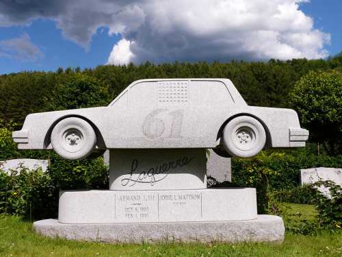 A granite monument shaped like a car, honoring Armand L. Lijh and Lionel Mattson, with a cloudy sky in the background.