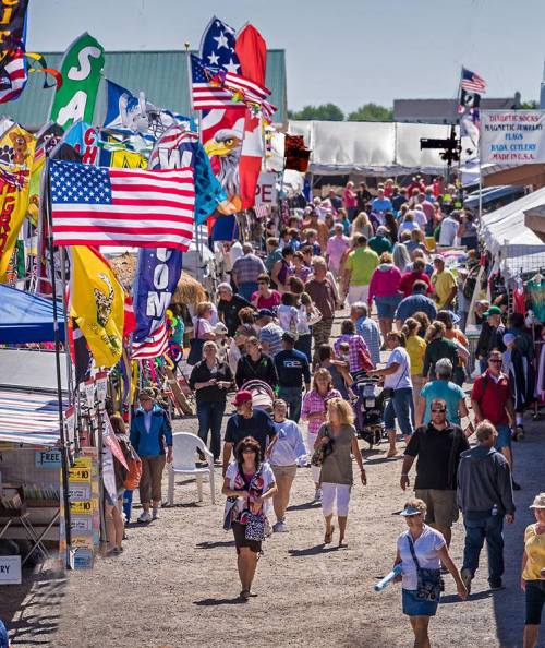A bustling outdoor market filled with people, colorful flags, and various stalls under a clear blue sky.