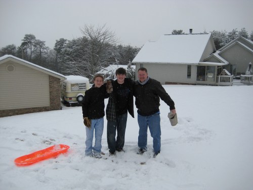 Three people stand in the snow, smiling, with a bright orange sled nearby and houses in the background.