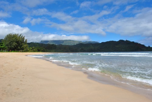 A serene beach with gentle waves, lush green mountains in the background, and a partly cloudy blue sky.
