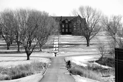 A black and white image of a long driveway leading to a large, old house surrounded by bare trees and open fields.