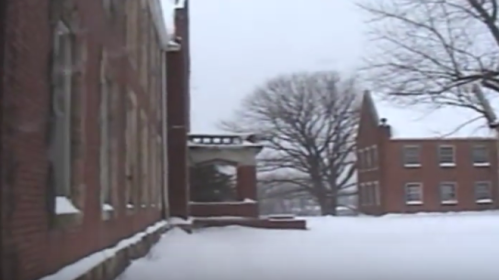 Snow-covered landscape featuring brick buildings and bare trees in a winter setting.