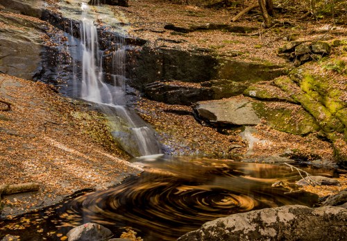 A serene waterfall cascades over rocks, surrounded by autumn leaves and a swirling pool of water below.