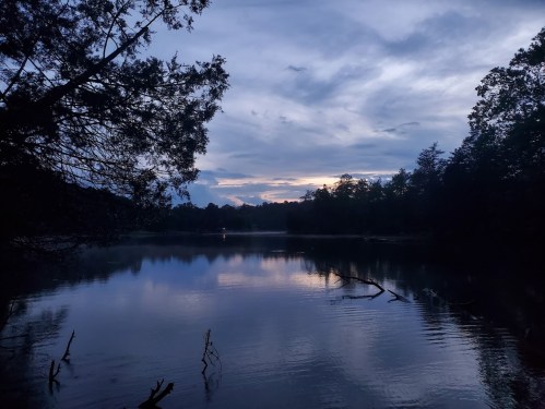 A serene lake at dusk, reflecting clouds and trees, with a hint of sunset colors in the sky.