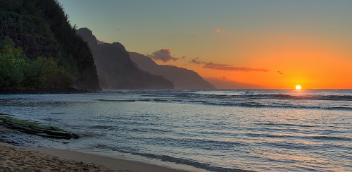 Sunset over a tranquil beach, with mountains in the background and gentle waves lapping at the shore.