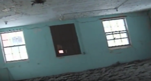 An empty, dilapidated room with peeling paint, two windows, and a dark opening on one wall.