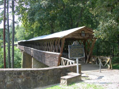 A wooden covered bridge with a metal roof, surrounded by trees and a historical marker nearby.