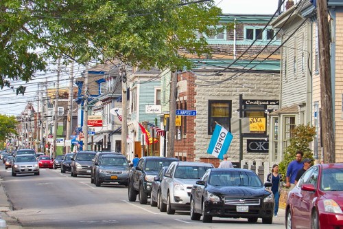 A busy street lined with shops and parked cars, featuring pedestrians walking along the sidewalk under a clear sky.