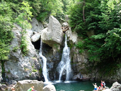 A serene waterfall cascades over rocks into a clear pool, surrounded by lush greenery and people enjoying the scenery.