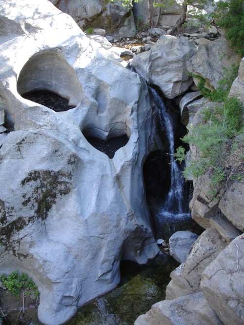 A waterfall cascades over smooth, rocky formations with natural pools and greenery surrounding the scene.