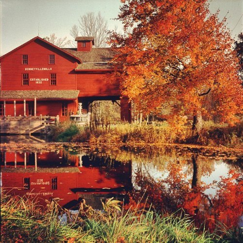 A red mill building surrounded by vibrant autumn foliage, reflecting in a calm pond.