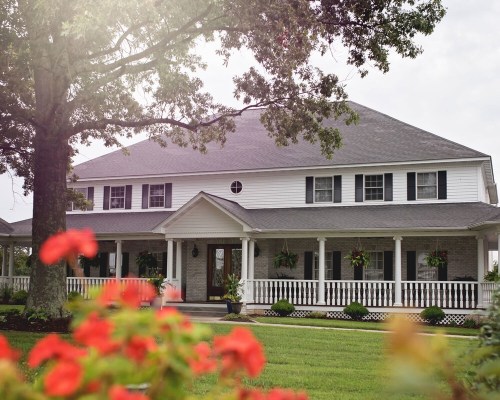 A charming two-story house with a large porch, surrounded by greenery and colorful flowers in the foreground.