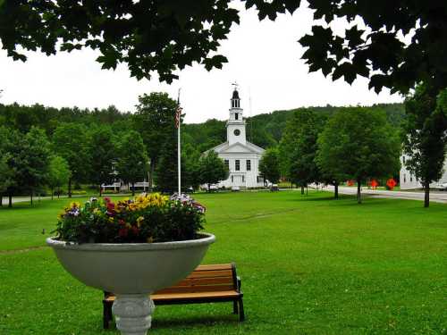 A green park with a flower planter, bench, and a white church in the background surrounded by trees.