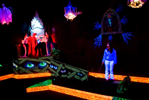 A dark, glowing mini-golf scene with spooky decorations, including ghosts, spider webs, and eerie eyes.