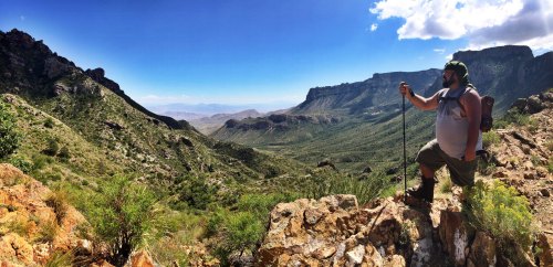 A hiker stands on a rocky outcrop, overlooking a vast green valley and mountains under a clear blue sky.