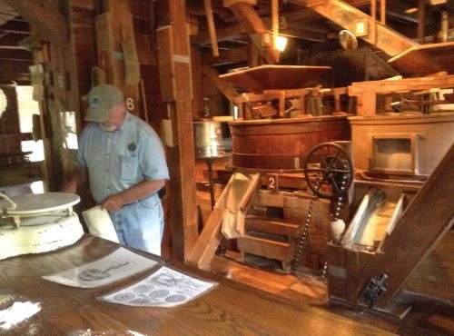 A man in a blue shirt works in a historic mill, surrounded by wooden machinery and milling equipment.