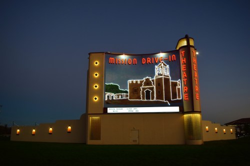 A brightly lit vintage drive-in theater sign at dusk, featuring "Mission Drive-In Theatre" in neon lights.