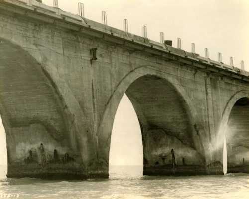 Black and white photo of a concrete bridge with arches over water, showcasing weathered structures and supports.