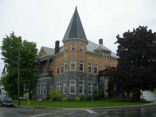 Historic building with a pointed roof, brick and stone exterior, surrounded by trees and a grassy area.