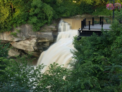 A waterfall cascades over rocky cliffs, surrounded by lush greenery and a viewing platform nearby.