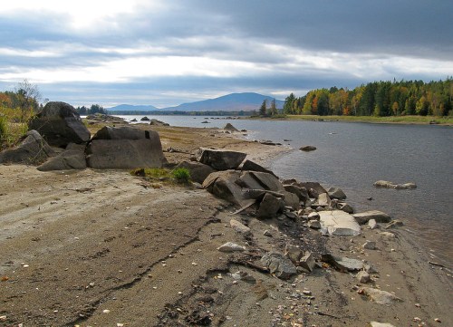 A serene riverbank scene with rocky shores, gentle waves, and distant mountains under a cloudy sky.