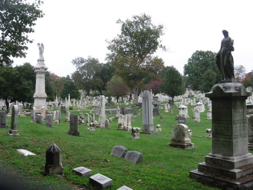 A serene cemetery with various gravestones and statues, surrounded by trees under a cloudy sky.