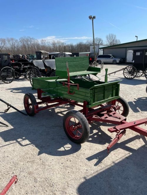 A green wooden cart with red wheels, parked on gravel, surrounded by other horse-drawn vehicles in a rural setting.